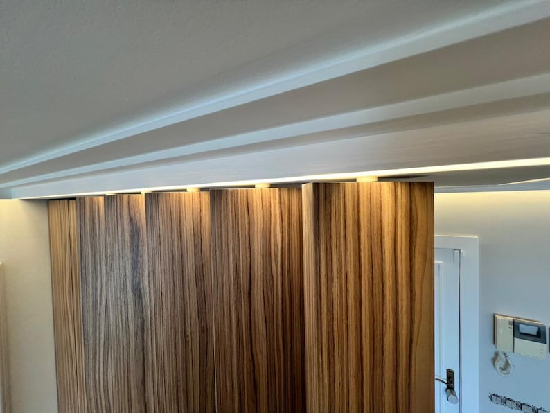 May include: A close-up of a wooden sliding door with vertical grain patterns. The door is a warm brown color with lighter streaks. The ceiling above has white beams and recessed lighting. The door is partially open, revealing a white door frame and wall.