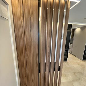 May include: Interior view of a modern space featuring a wooden cabinet with vertical grain and a slatted wooden room divider. The wood has a warm, natural tone. The floor is tiled with a light-colored stone.