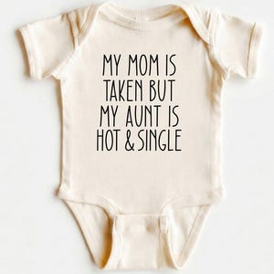 May include: Cream-colored baby onesie with the text "MY MOM IS TAKEN BUT MY AUNT IS HOT & SINGLE" in black. The onesie has short sleeves and a snap closure at the bottom. The text is centered on the front of the garment.