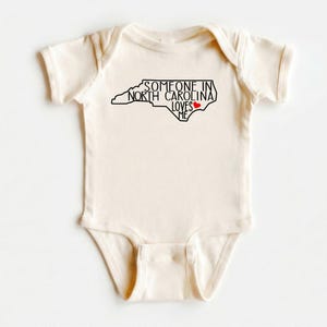 Body para bebé con el mensaje "Alguien en Carolina del Norte me ama", camiseta adorable para bebé, regalos para baby shower, camiseta para bebé con el mensaje "Amado", body para bebé con el estado, camiseta natural para bebé. imagen 1
