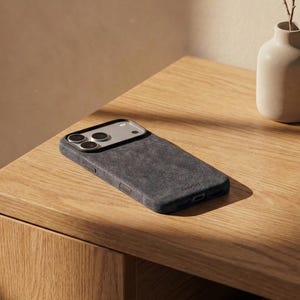 May include: A dark grey phone case with a textured finish lies on a wooden table. The case features a camera aperture and a streamlined design. A small, beige vase with a sprig of dried flowers is also present.