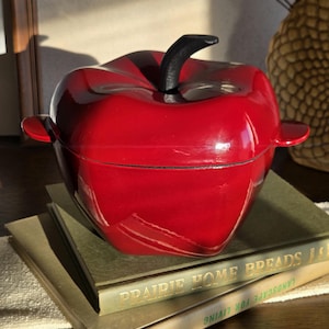 May include: A glossy red apple-shaped casserole dish with a black handle and side handles. The dish sits atop a stack of vintage books with gold lettering. The overall theme is kitchenware and home decor.