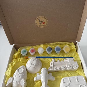 May include: A craft kit in a cardboard box containing five white ceramic figures shaped like a rocket, a globe, a train, an aeroplane, and a bus. The kit includes a blue paintbrush and a strip of six small paint pots. A sticker reads Thank you.
