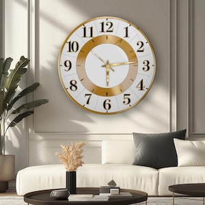 May include: A large, round wall clock with a white face, gold-colored hands, and black numerals. The clock has a gold-colored frame and a decorative gold-colored inner ring. The clock is mounted on a light-colored wall.