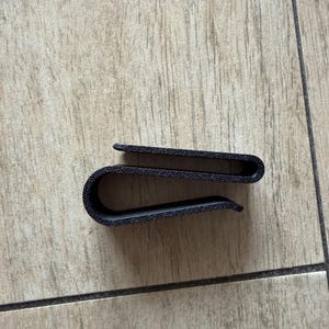 May include: A black, U-shaped money clip. The clip is made of a textured material and is designed to hold folded banknotes securely. The design is simple and functional, suitable for everyday use.