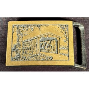 May include: A rectangular brass belt buckle featuring a raised, textured relief of a covered bridge surrounded by trees. The metal surface has a brushed gold finish, while the recessed areas of the bridge scene are darkened to provide contrast.