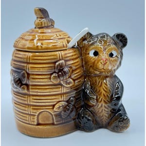 May include: A ceramic honey pot shaped like a beehive with a bear cub figurine. The honey pot is brown with a lid and a white spoon. The bear cub is brown and black with a smiling face. The beehive has bee decorations.