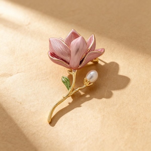 May include: A pink and gold floral brooch in the shape of a magnolia flower. The brooch has a pearl accent and green leaves. The petals are pink with gold trim, and the stem is gold. A lovely accessory for any outfit.