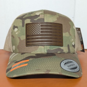 May include: A camouflage baseball cap with a brown leather American flag patch. The hat has a mesh back and a curved brim. The front of the hat has the word "MULTICAM" printed on it.