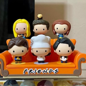May include: A set of Friends character figurines, each with a distinctive hairstyle and outfit, arranged on an orange sofa-shaped base. The base is inscribed with the show's logo. The figurines are in a cartoon style.