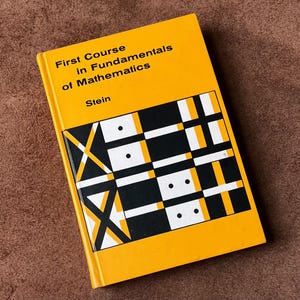 Puede incluir: Un libro amarillo titulado "First Course in Fundamentals of Mathematics" de Stein. La portada presenta un diseño geométrico en negro, blanco y naranja. El libro está sobre una superficie marrón.