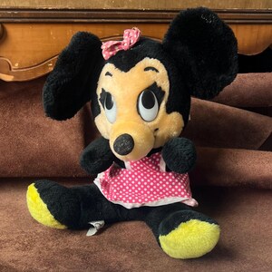 Pu&ograve; includere: Un peluche di Minnie Mouse con pelliccia nera, grandi orecchie nere e un viso giallo. Indossa un vestito rosa a pois e un fiocco rosa. La bambola ha i piedi gialli e grandi occhi espressivi.
