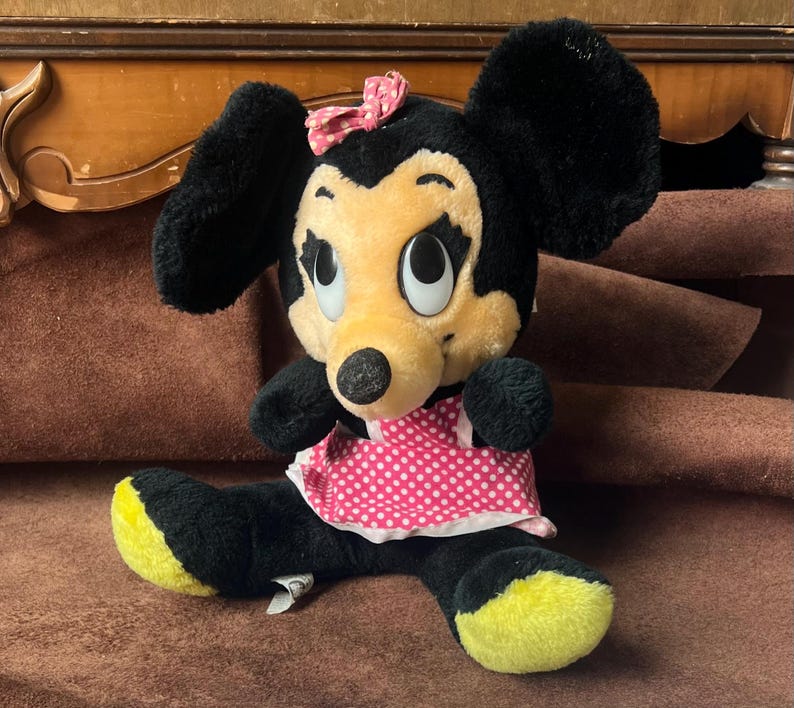 Pu&ograve; includere: Un peluche di Minnie Mouse con pelliccia nera, grandi orecchie nere e un viso giallo. Indossa un vestito rosa a pois e un fiocco rosa. La bambola ha i piedi gialli e grandi occhi espressivi.