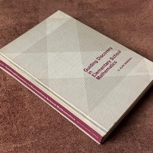 Può includere: Un libro con copertina rigida intitolato "Guiding Discovery in Elementary School Mathematics" con un motivo geometrico sulla copertina. Il dorso e i bordi sono di colore viola scuro. Il libro è su una superficie marrone.