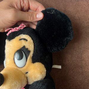Pu&ograve; includere: Una bambola di peluche di Minnie Mouse con pelliccia nera, un fiocco rosa a pois bianchi e un viso color camoscio. La bambola ha grandi occhi espressivi e un aspetto morbido e coccoloso. L'orecchio &egrave; tenuto da una mano.