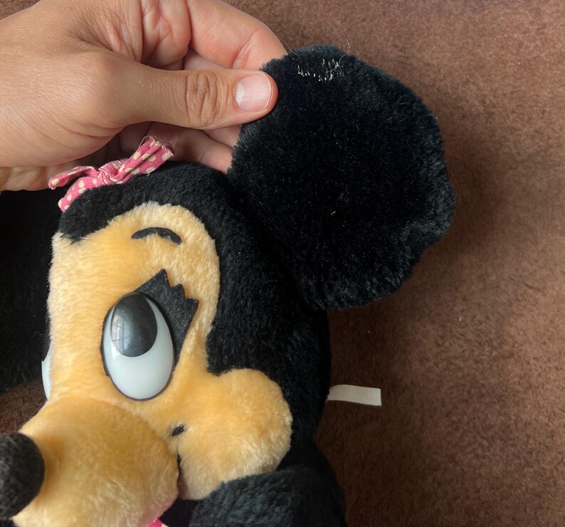 Pu&ograve; includere: Una bambola di peluche di Minnie Mouse con pelliccia nera, un fiocco rosa a pois bianchi e un viso color camoscio. La bambola ha grandi occhi espressivi e un aspetto morbido e coccoloso. L'orecchio &egrave; tenuto da una mano.