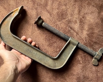May include: An antique C-clamp, 8 inches in size, held in a hand. The clamp is made of aged metal with a threaded rod and a tightening handle. The text "ADY CLAMP 8" is visible on the clamp.