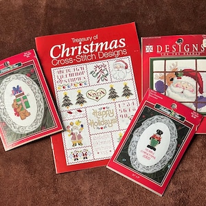 Puede incluir: Una colección de libros y kits de diseños de punto de cruz navideños. Los libros tienen cubiertas rojas con títulos como "Treasury of Christmas Cross-Stitch Designs". Los kits presentan patrones de punto de cruz festivos.