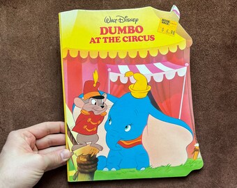 May include: A vintage Walt Disney book cover featuring Dumbo the elephant with Timothy Q. Mouse. The book is titled "Dumbo at the Circus" and has a red, white, and pink circus tent backdrop. The book cover has a price tag of $4.98.