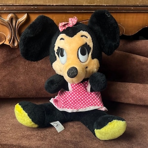Pu&ograve; includere: Una bambola di peluche di Minnie Mouse con pelliccia nera, viso giallo e vestito a pois rosa. La bambola ha un fiocco rosa e piedi gialli. Un'etichetta &egrave; attaccata alla bambola.