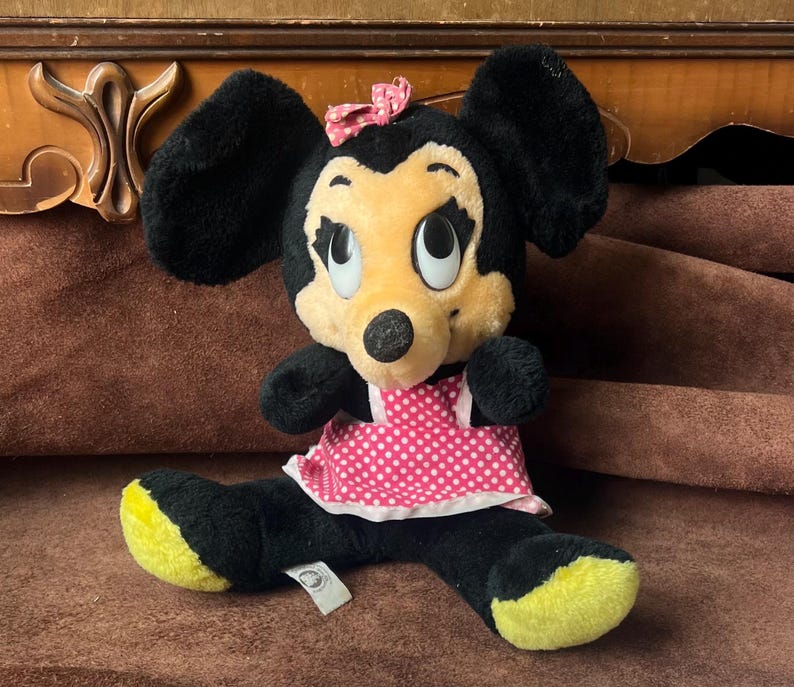 Pu&ograve; includere: Una bambola di peluche di Minnie Mouse con pelliccia nera, viso giallo e vestito a pois rosa. La bambola ha un fiocco rosa e piedi gialli. Un'etichetta &egrave; attaccata alla bambola.