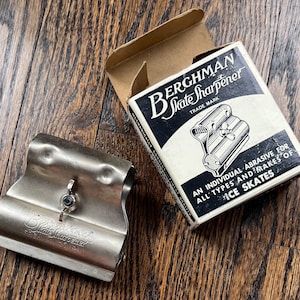 Pode incluir: Um afiaador de patins vintage de metal prateado na sua caixa original. A caixa é branca com letras pretas e um desenho do afiaador. A caixa diz "Berchman Skate Sharpener Trade Mark" e "Um abrasivo individual para todos os tipos e marcas de patins de gelo."