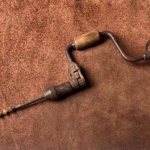 May include: A vintage hand brace drill featuring a dark metal frame, a rotating wooden handle, and a wooden head knob. The tool includes a metal chuck mechanism and a long, spiraled auger bit extending from the front, showing signs of age.
