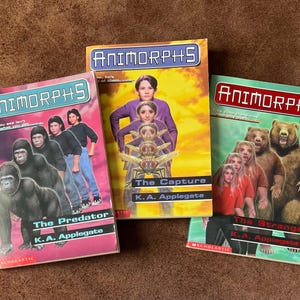 Puede incluir: Se muestran tres libros de bolsillo de la serie Animorphs. Las portadas presentan ilustraciones de personas transformándose en animales, con títulos como "The Predator" y "The Capture". Los libros están sobre un fondo marrón.