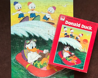 May include: A 100-piece jigsaw puzzle featuring Donald Duck and his nephews on surfboards. The box is red with a white border and the words "Jigsaw Puzzle" and "Donald Duck" in white text. The puzzle is 14" x 18" or 35.5 cm x 45.7 cm.
