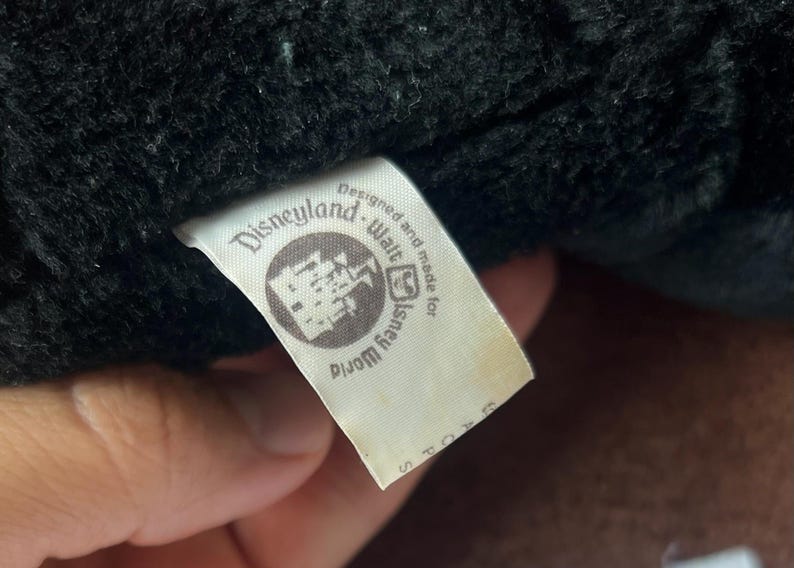 Pu&ograve; includere: Primo piano di un'etichetta bianca con testo nero, attaccata a un tessuto nero. L'etichetta recita "Designed and made for Disneyland Walt Disney World Stores". L'etichetta ha un logo circolare.