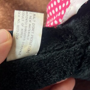 Pu&ograve; includere: Primo piano di un giocattolo di peluche nero con un fiocco rosa e bianco a pois. Un'etichetta bianca con testo nero recita "WALT DISNEY PRODUCTIONS" e altri dettagli sul giocattolo. Il giocattolo &egrave; fatto di nuovi materiali e ritagli sminuzzati.