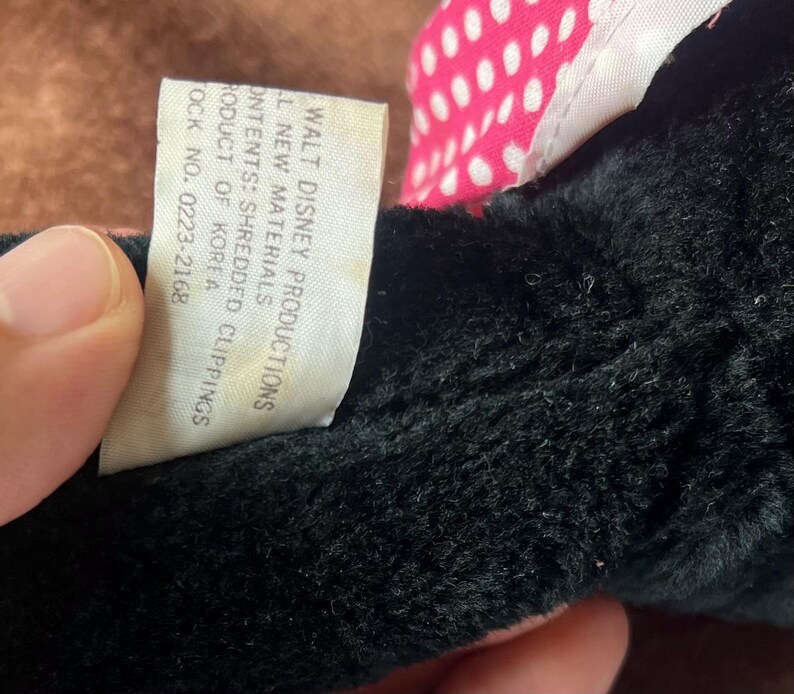 Pu&ograve; includere: Primo piano di un giocattolo di peluche nero con un fiocco rosa e bianco a pois. Un'etichetta bianca con testo nero recita "WALT DISNEY PRODUCTIONS" e altri dettagli sul giocattolo. Il giocattolo &egrave; fatto di nuovi materiali e ritagli sminuzzati.