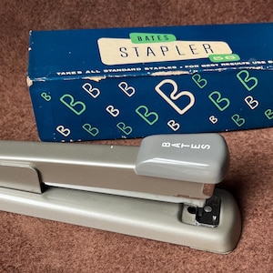 May include: A silver Bates stapler with a gray top and the word "Bates" printed on it. The stapler is sitting on a brown surface. The box for the stapler is in the background. The box is blue with white letters and a pattern of green "B"s.