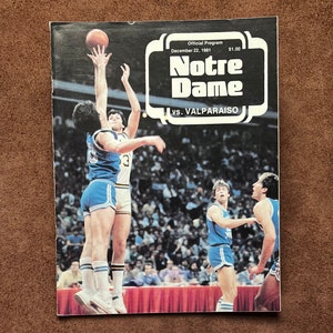 Può includere: Un programma di gioco di basket vintage di Notre Dame del 22 dicembre 1981, con il logo della squadra e le parole "Notre Dame vs. Valparaiso". La copertina del programma mostra un giocatore di basket con una maglia blu che salta per tirare a canestro.
