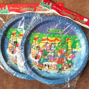 Puede incluir: Dos bandejas de servir navideñas azules y blancas con una escena de invierno nevada y un escaparate de tienda de juguetes. El escaparate tiene la palabra "TOYS" encima. Las bandejas están envueltas en plástico.