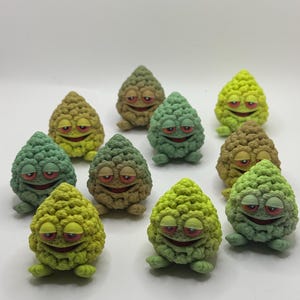Op de afbeelding: Een verzameling grillige, kegelvormige beeldjes met lachende gezichten en kleine voetjes. De beeldjes zijn in verschillende tinten groen, geel en bruin, met rode lippen en gele ogen. Ze lijken handgemaakt en hebben een textuur.