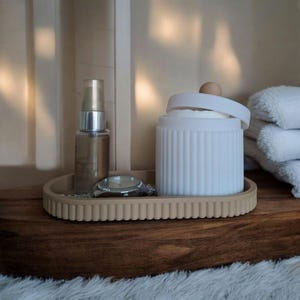 Könnte beinhalten: Ein beigefarbener, geriffelter Tablett hält eine silberne Uhr, eine Hautpflegeflasche und ein weißes, geripptes Glas mit einem Holzknauf. Das Tablett steht auf einer dunklen Holzoberfläche, mit einem Stapel weißer Handtücher im Hintergrund.