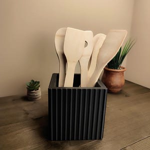 May include: A black, ribbed utensil holder filled with wooden cooking spoons and spatulas. The holder sits on a wooden surface, with small potted plants in the background. The utensils are made of light-coloured wood.