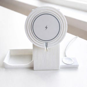 Peut inclure: Station de recharge sans fil blanche avec un socle circulaire portant un symbole d'éclair. La station comprend un support, un petit plateau et une base de recharge séparée pour une montre connectée. Design minimaliste.