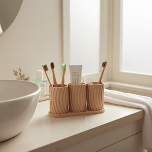 Op de afbeelding: Een set van drie lichtbruine, geribbelde tandenborstelhouders op een bijpassende tray, met tandenborstels en een tube tandpasta. De tandenborstels hebben bruine en groene borstelharen. De scène speelt zich af in een badkamer.