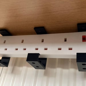 Puede incluir: Una regleta blanca con múltiples enchufes y una luz indicadora roja está montada debajo de una superficie de madera. Los soportes negros aseguran la regleta. Un cable de alimentación blanco se extiende desde la regleta.