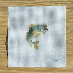 May include: A square needlepoint canvas featuring a jumping fish design. The fish is primarily green and yellow, with a dark purple mouth. The word "SALT" is stitched in blue in the lower right corner. The canvas is on a light blue background.