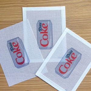 May include: Three needlepoint canvases featuring a Diet Coke can design. The cans are light blue with red text and are set against a white mesh background. The canvases are arranged on a light brown surface.