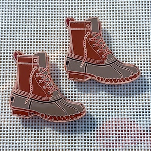 May include: Two enamel pins in the shape of brown and gray duck boots. The boots have red laces and a decorative border. The pins are on a white grid background.