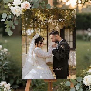 May include: A rectangular wedding welcome sign featuring a photograph of a couple, with the word Welcome in white script at the top and the names LYDIA & NATHAN with the date MARCH 28, 20XX printed in gold lettering at the bottom of the image.