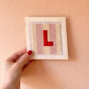 Puede incluir: Una pieza de punto de cruz hecha a mano con la letra "L" en rojo, sobre un fondo de rayas rosas y amarillas. El bordado est&aacute; montado sobre un lienzo cuadrado, perfecto para decoraci&oacute;n personalizada.