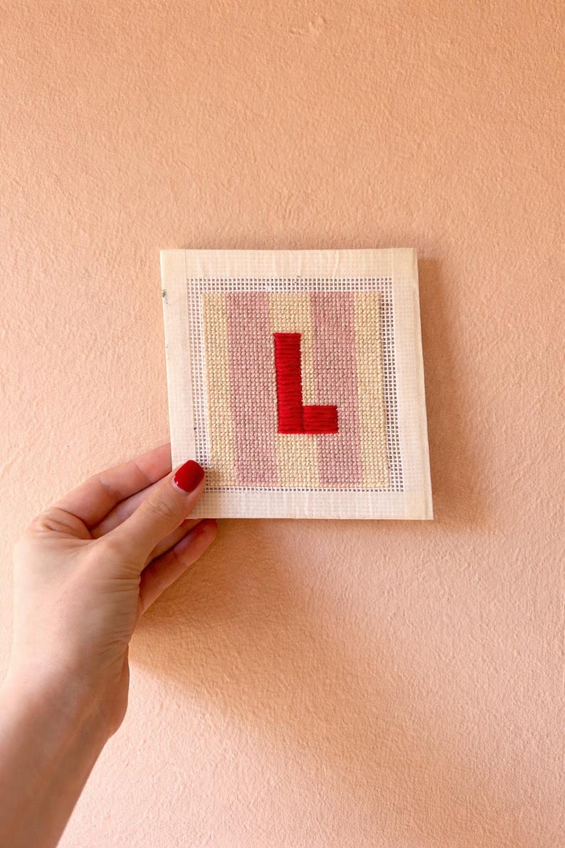 Puede incluir: Una pieza de punto de cruz hecha a mano con la letra "L" en rojo, sobre un fondo de rayas rosas y amarillas. El bordado est&aacute; montado sobre un lienzo cuadrado, perfecto para decoraci&oacute;n personalizada.