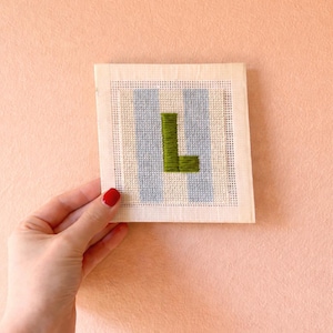 Puede incluir: Un bordado a mano con la letra "L" en verde oliva, sobre un fondo de rayas verticales azul claro. La pieza se muestra sobre un lienzo color crema, creando una est&eacute;tica vintage. El fondo es de color melocot&oacute;n s&oacute;lido.