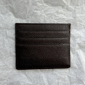 May include: A dark brown leather card holder with multiple card slots. The card holder has a textured surface and is rectangular in shape. The background is a crumpled white paper.