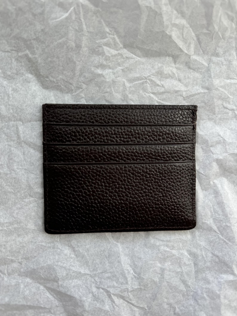 May include: A dark brown leather card holder with multiple card slots. The card holder has a textured surface and is rectangular in shape. The background is a crumpled white paper.